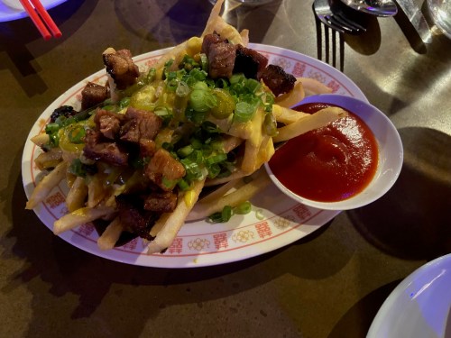 A plate of loaded fries topped with green onions and chunks of meat, served with a side of ketchup.