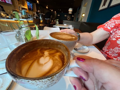 Two hands holding coffee cups with latte art, set on a table in a cozy café with a blurred background.