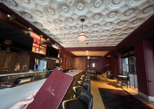 A cozy bar interior with a decorative ceiling, empty tables, and a person holding a menu in the foreground.