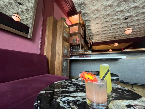 A cozy bar scene featuring two colorful cocktails on a marble table, with plush seating and a stylish ceiling.