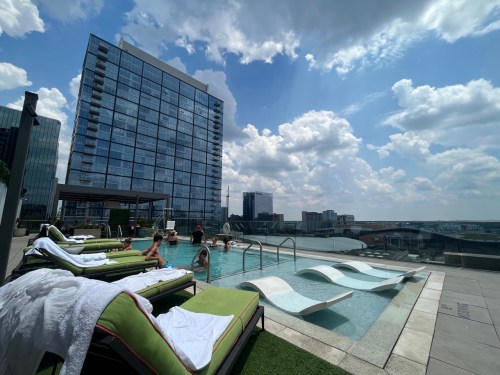 Rooftop pool with lounge chairs, people swimming, and a city skyline under a partly cloudy sky.