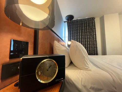 A bedside clock on a wooden table next to a neatly made bed with white pillows and patterned curtains in the background.