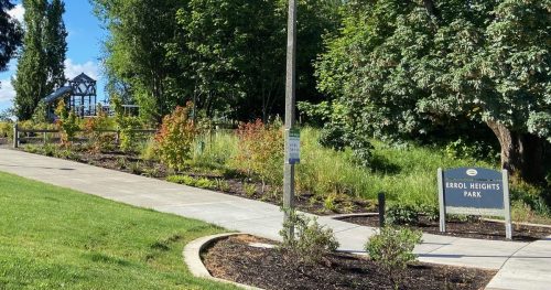A paved path leads through a park with trees and greenery under a blue sky, featuring a sign for "Lions Park."