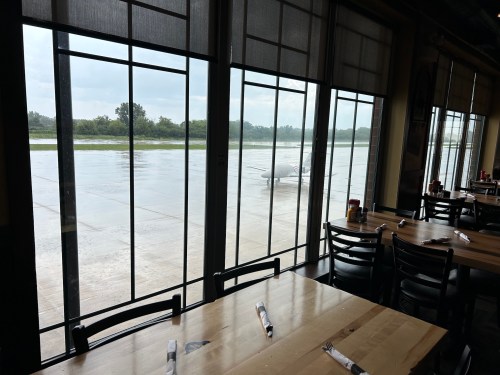 View from a restaurant with wooden tables, looking out at a rainy airport runway and a small airplane.