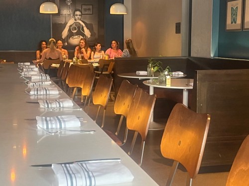 A modern restaurant interior with a long table set for dining and a group of people seated in the background.