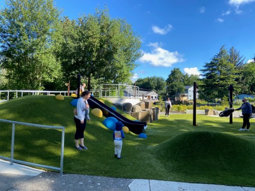 A playground with slides and grassy hills, where a child walks towards an adult, with people enjoying the space.