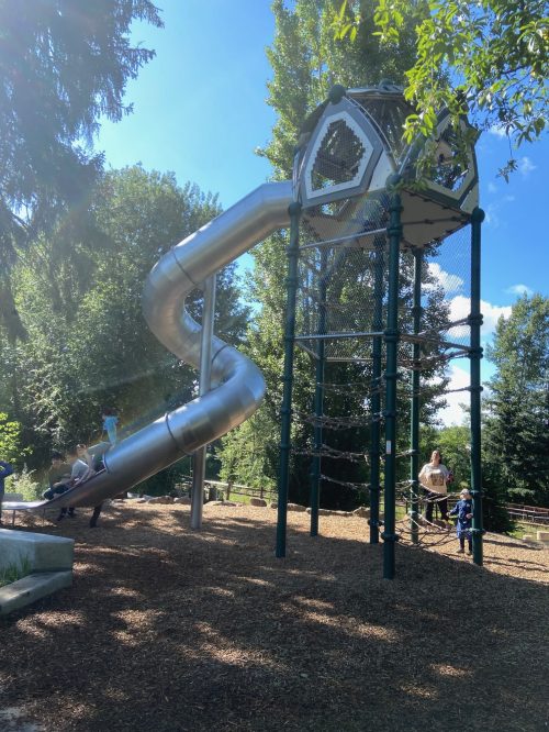 A large metal slide spirals down from a tall play structure in a sunny park, with children playing nearby.