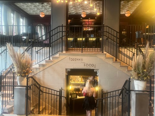 A woman in a black dress walks down stairs to a bar area, with decorative plants and warm lighting in a stylish venue.