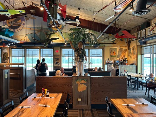 A lively restaurant interior with vintage airplane decor, wooden tables, and a statue of a boy holding a toy plane.