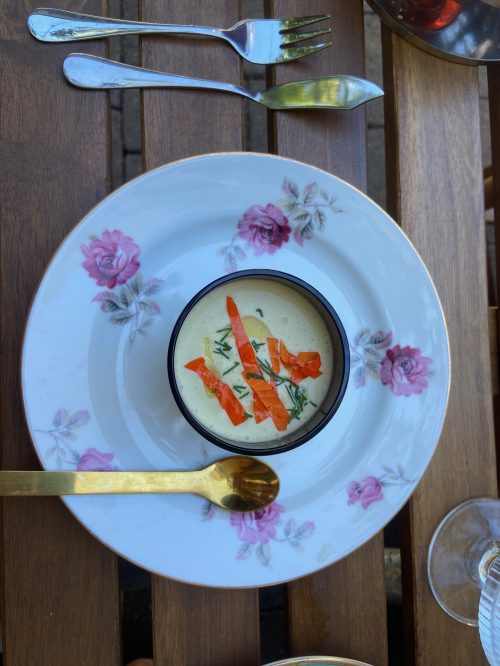 A bowl of creamy soup garnished with orange strips, served on a floral plate, with cutlery on a wooden table.