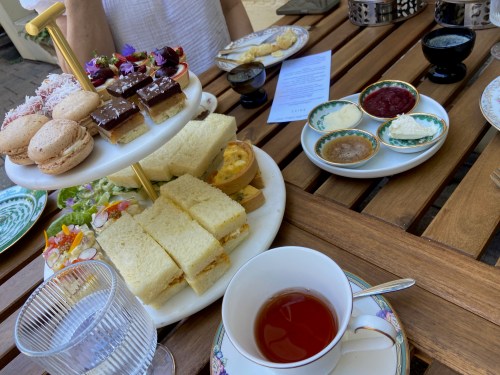 A tiered platter of sandwiches, pastries, and desserts, with a cup of tea on a wooden table.