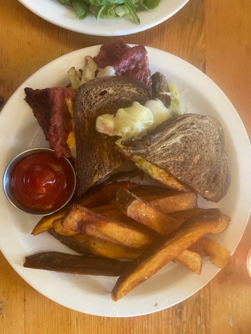 A plate with a sandwich made of dark bread, melted cheese, and lettuce, served with sweet potato fries and ketchup.