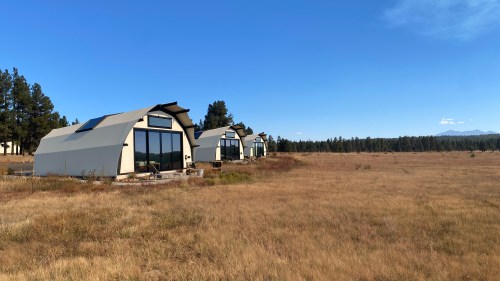 Three modern, tent-like cabins in a grassy field, surrounded by trees under a clear blue sky.