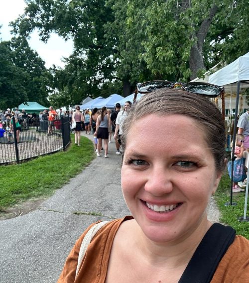 A smiling person with sunglasses stands on a path at a busy outdoor event with tents and people in the background.