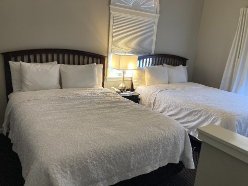 A cozy bedroom with two beds, white bedding, a bedside lamp, and natural light from a window.
