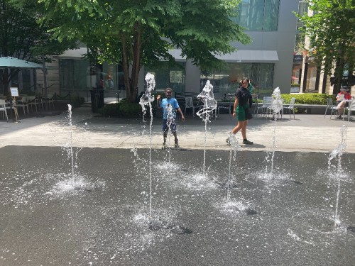 A child plays in a splash pad while an adult walks nearby, surrounded by trees and outdoor seating.