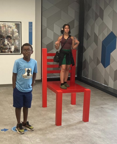 A girl stands on a large red chair while a boy poses beside it in a modern indoor space.