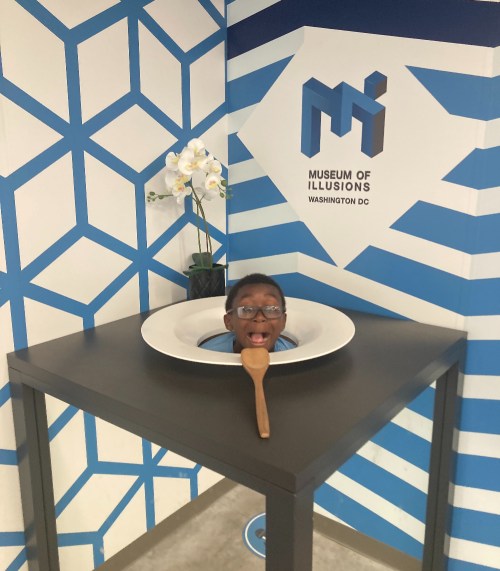 A child peeks out from a large plate on a table at the Museum of Illusions in Washington, DC, with a wooden spoon nearby.