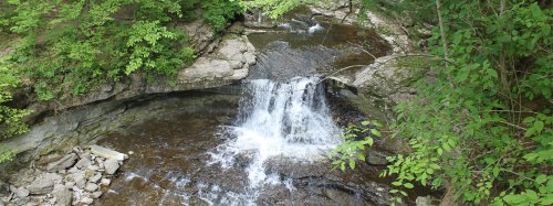 A serene waterfall cascading over rocks, surrounded by lush green foliage and a tranquil stream below.