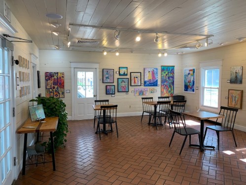 A bright art gallery with colorful paintings on the walls, tables, and chairs arranged for visitors.