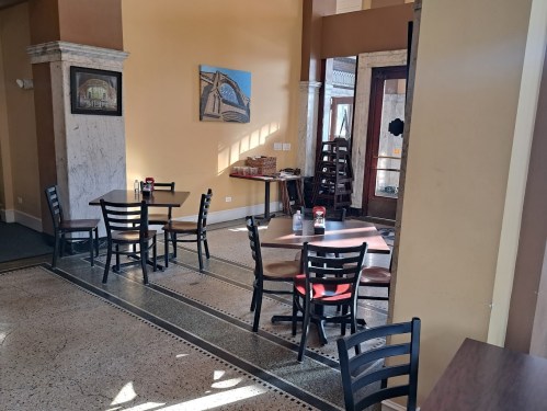 A cozy restaurant interior with empty tables, marble columns, and a painting on the wall. Natural light fills the space.