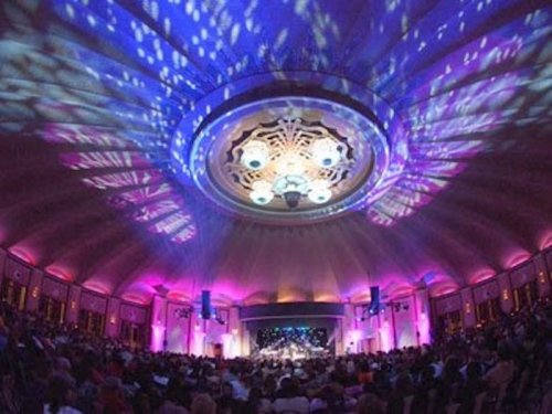 A vibrant concert hall filled with an audience, featuring colorful lights and an ornate ceiling design.