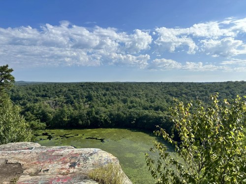 A scenic view from a rocky overlook, featuring lush green trees and a pond under a blue sky with scattered clouds.