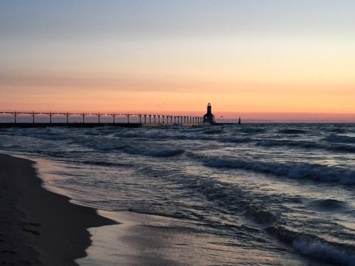 A serene beach scene at sunset, featuring a pier and lighthouse silhouetted against a colorful sky. Waves gently lap the shore.