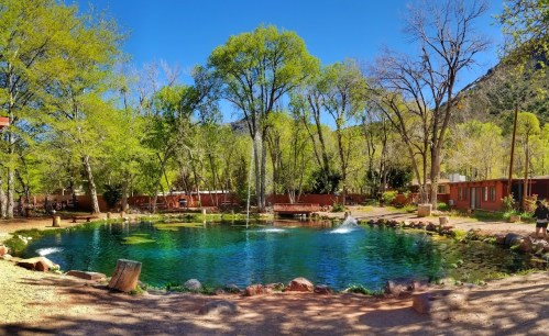 A serene pond surrounded by lush green trees and rustic buildings under a clear blue sky.