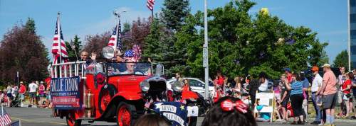 A vintage red car in a parade, decorated with American flags and carrying people celebrating a festive event.