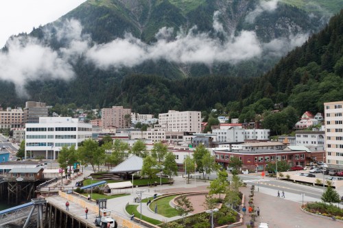 A scenic view of a coastal town surrounded by mountains, with buildings, trees, and people walking along a pathway.