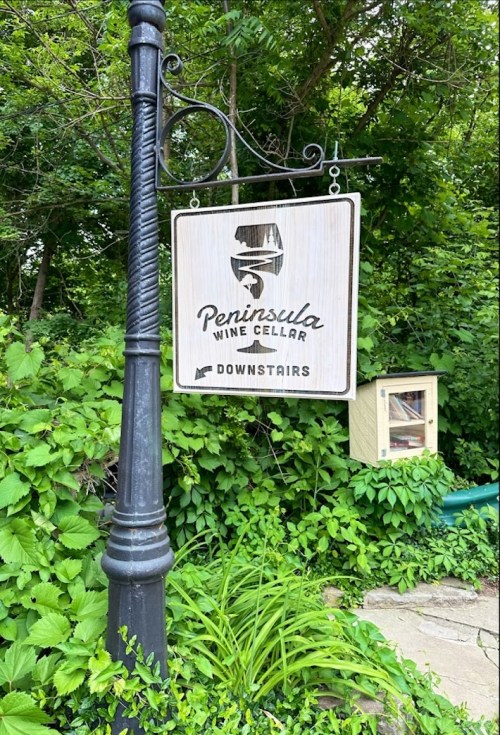 Sign for "Peninsula Wine Cellar" hanging from a post, surrounded by lush greenery, indicating "Downstairs."