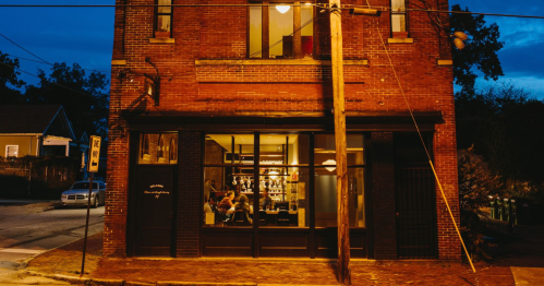 A cozy brick building at dusk, with warm lights glowing from the windows and people inside.