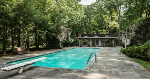 A serene outdoor pool surrounded by lush greenery and stone walls, with a diving board and a modern building nearby.