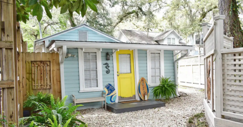 A charming blue cottage with a yellow door, surrounded by greenery and decorative surfboards on the porch.