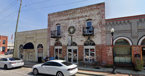 Historic brick building with large windows, a clock, and outdoor seating, set in a small town street.