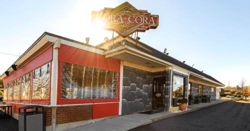 Exterior of a restaurant named "Cora Cora," featuring large windows and a bright sun flare in the background.