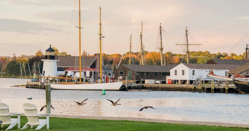 A serene harbor scene with a lighthouse, sailboats, and seagulls flying over calm waters at sunset.