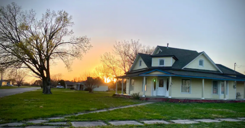 A charming yellow house with a porch, surrounded by grass, at sunset with trees silhouetted against the colorful sky.