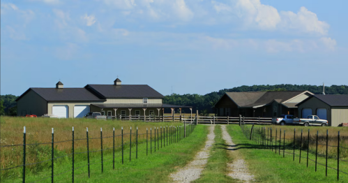A gravel driveway leads to two farm buildings surrounded by green fields and a blue sky with scattered clouds.