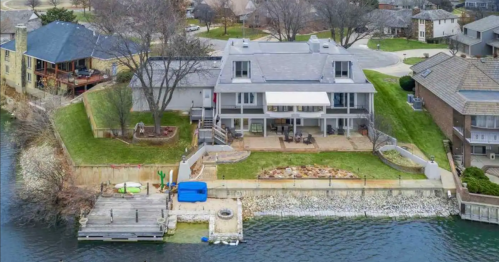 Aerial view of a large waterfront home with a patio, dock, and landscaped yard by a calm lake.
