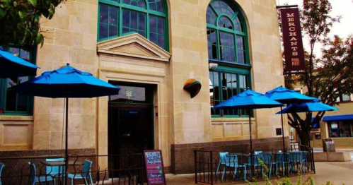 Exterior of a restaurant named "Merchants," featuring blue umbrellas and outdoor seating.