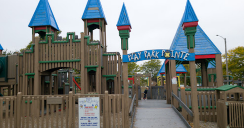 Colorful playground with blue-roofed structures and a sign reading "Play Park Pointe." Trees in the background.