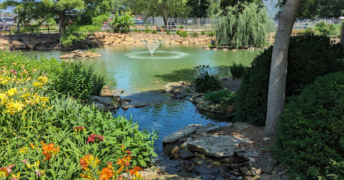 A serene pond surrounded by lush greenery and colorful flowers, featuring a fountain in the center.