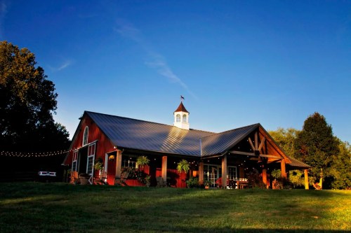 A red barn-style building with a cupola, surrounded by greenery and string lights, under a clear blue sky.