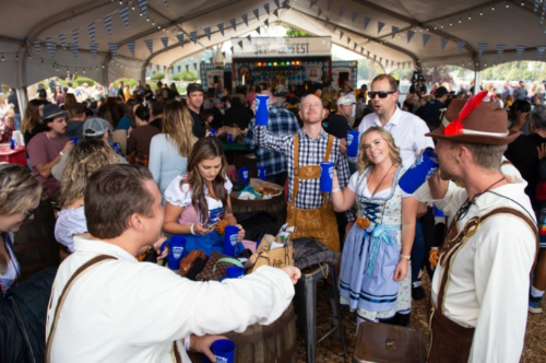 A lively festival scene with people in traditional attire raising blue cups under a large tent filled with guests.