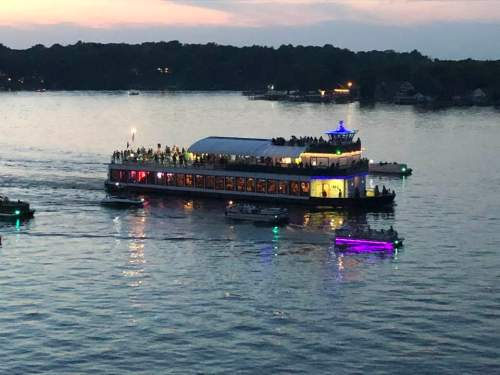 A brightly lit boat with people on deck sails on a calm lake at sunset, surrounded by smaller boats.