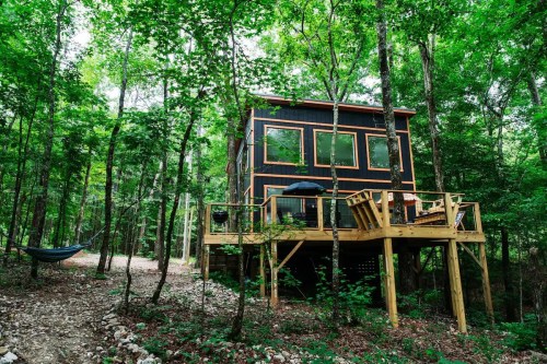 A modern cabin on stilts surrounded by lush green trees, with a deck and hammock nearby.