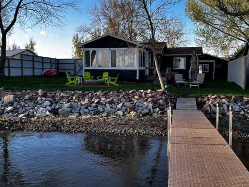A cozy lakeside cabin with green chairs, a dock, and a rocky shoreline under a clear blue sky.