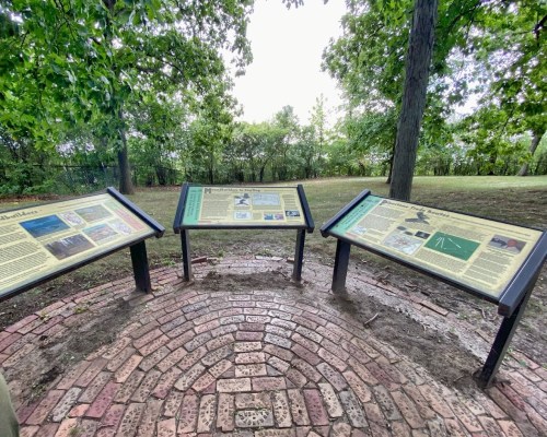 Three informational panels in a circular arrangement on a brick path, surrounded by trees and greenery.